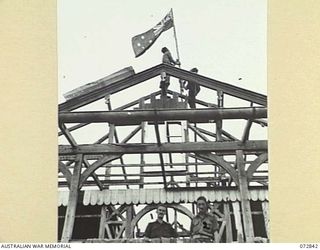 ALEXISHAFEN, NEW GUINEA. 1944-04-30. MEMBERS OF THE 30TH INFANTRY BATTALION MOUNT THE AUSTRALIAN FLAG WHICH WAS PRESENTED TO THE BATTALION BY THE CREW OF HMAS BUNDABERG, THE RUINS OF FORMER LIVING ..