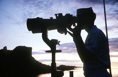 A sailor aboard the tank landing ship USS RACINE (LST 1191) studies his surroundings with the ship's binoculars at dusk. The vessel is in the region as part of a civic action program