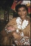 New mother, holding newborn, wears long fiber shawl, flower garland, and skirt valuable on head, and betel nut paste on face