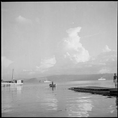 Man standing in a rowboat and another standing on a boat ramp with HMAS Australia [?] in the background, Rabaul Harbour, New Guinea, 1937 / Sarah Chinnery