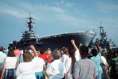 Crew members man the rails aboard the amphibious assault ship USS GUAM (LPH-9) as family and friends wave goodbye from the pier. The GUAM is departing for the Middle East in support of Operation Desert Shield