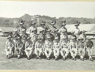 Outdoor group portrait of members of the 18th Field Ambulance on the unit parade ground with flagpole and Regimental Aid Post in the background. Identified in the back row from left to right: ..