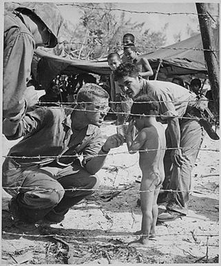 Marines try to soothe a crying child by offering a shiny rations tin. Children are sheltered with their families in a camp set up for refugees from battle areas by U.S. Marine Civil Affairs authorities on Saipan.