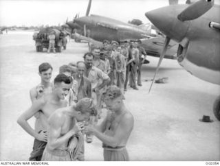 KIRIWINA, TROBRIAND ISLANDS, PAPUA. 1944-02-15. MATCHES ARE SCARCE IN NEW GUINEA JUNGLES AND AT "SMOKO" TIME AIRMEN OF NO. 76 (KITTYHAWK) SQUADRON RAAF LINE UP IN FRONT OF THE AIRCRAFT TO GET A ..