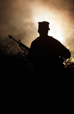 A Marine is silhouetted against a buring flare as he moves toward a Seabee camp during Exercise Kennel Bear '89. Marines from Marine Barracks Guam are acting as aggressors against Seabees from Naval Mobile Construction Battalion 3 (NMCB-3) and Marines from the 3rd Force Service Support Group (3RD FSSG)