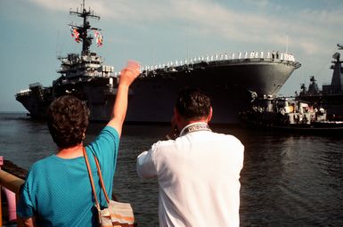 A mother waves from the pier as her son departs for the Persian Gulf aboard the amphibious assault ship USS GUAM (LPH-9), one of several ships deploying from Norfolk in response to Iraq's invasion of Kuwait. The large harbor tugs WATHENA (YTB-825) and ANOKE (YTB-810) are maneuvering the vessel away from the pier