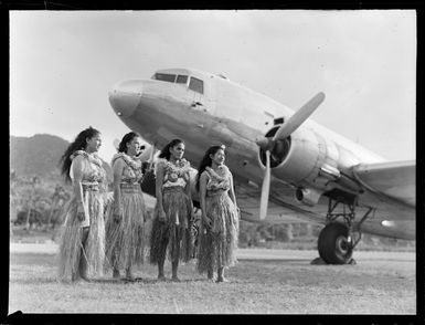 Four unidentified local girls wearing hula skirts in front of a C47 transport aircraft, Rarotonga airfield, Cook Islands