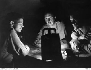 LAE, NEW GUINEA, 1943-10-13. SITTING AROUND A HURRICANE LAMP IN THE DOCK AREA ARE, LEFT TO RIGHT:- SX2663 SERGEANT E.E. SMITH; VX72048 SERGEANT F. MCKECKNIE; SX13471 SERGEANT G.R. MAINWARING, ALL ..