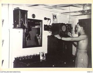 SIMPSON HARBOUR, NEW BRITAIN. 1945-10-13. SUB-LIEUTENANT M.R. BUTLER, ROYAL AUSTRALIAN NAVY STANDS AT THE BAR IN THE WARD ROOM OF THE MOTOR LAUNCH 825 AND TUNES IN THE RADIO. THE VESSEL IS THE ONLY ..
