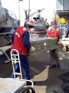 United States Navy, Aviation Ordnancemen move weapons, in a shipping container, from one of the ship's elevators to a waiting AH-1 "Sea Cobra" attack helicopter. A UH-1N "Huey" is in background. On board the USS GUAM (LPH 9) an amphibious assault ship currently in the Persian Gulf as part of an increased military presence to enforce U.N. sanctions against Iraq