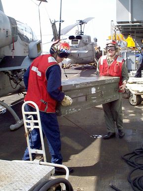 United States Navy, Aviation Ordnancemen move weapons, in a shipping container, from one of the ship's elevators to a waiting AH-1 "Sea Cobra" attack helicopter. A UH-1N "Huey" is in background. On board the USS GUAM (LPH 9) an amphibious assault ship currently in the Persian Gulf as part of an increased military presence to enforce U.N. sanctions against Iraq