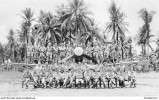 Large group portrait of pilots and administrative staff of No. 77 Squadron RAAF assembled on and in front of one of the squadron’s Curtiss P-40 Kittyhawk aircraft, at Gurney airstrip. Left to ..