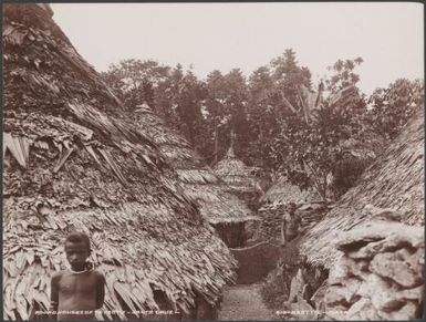 Two villagers standing between round houses of Te Motu, Santa Cruz Islands, 1906 / J.W. Beattie