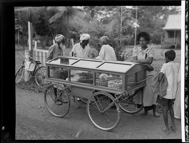 Unidentified Fijian men and woman beside a glass-sided cart displaying various foods, market at Ba, Fiji