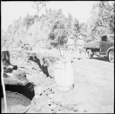 Two men and two trucks near a barrier made using casks to warn of erosion at the edge of the road, Rabaul, New Guinea, 1937 / Sarah Chinnery