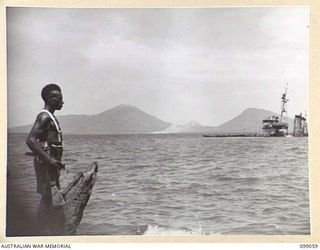 A LOCAL SOLDIER OF THE NEW GUINEA INFANTRY BATTALION, SURVEYS KARAVIA BAY SHOWING THE TWO VOLCANIC CRATERS, THE MOTHER MATUPI AND SOUTH DAUGHTER IN THE BACKGROUND. THE WRECKED JAPANESE YUGUMO CLASS ..