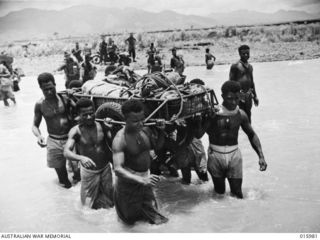 Ramu Valley, New Guinea. 21 October 1943. New Guinea natives from a forward dressing station carry supplies packed in a wire framed trailer across the Surinam River