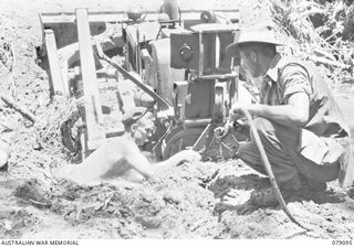 BOUGAINVILLE ISLAND. 1945-02-13. S52524 CORPORAL D.C. RUWOLDT (1) AND NX123140 LANCE CORPORAL F.J. NICOLLS (2) 61ST INFANTRY BATTALION, ATTACHING A TOW ROPE TO A BADLY BOGGED BULLDOZER ON A SECTION ..