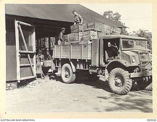 LAE, NEW GUINEA. 1944-12-12. DRIVER L.W. OSBORNE, ARMY CANTEENS SERVICE, (1), DIRECTING NATIVES SUPPLIED BY ANGAU IN THE UNLOADING OF BOTTLES AT A SOFT DRINK FACTORY WITHIN LAE BASE SUB-AREA