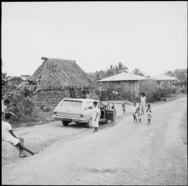Fijian family near a grass hut and houses, Lami, Fiji, 1966 / Michael Terry