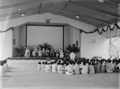 [A large group of pacific island children sitting on the floor in front of adults and nuns seated on a stage]