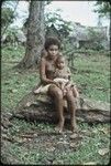 Adolescent girl, wearing shell necklace and dashes of betel nut paste on her cheeks, holds a baby on her lap