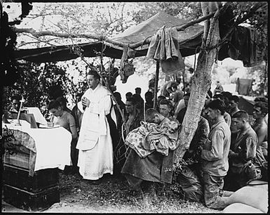 With a canvas tarpaulin for a church and packing cases for an altar, a Navy chaplain holds mass for Marines at Saipan. The service was held in memory of brave buddies who lost their lives in the initial landings.