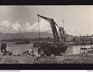 LAE, NEW GUINEA. 1943-10-12. THE WATERFRONT WITH AN AMPHIBIOUS DUKW IN THE FOREGROUND AND A FLOATING CRANE WORKING IN THE HARBOUR. AT LEFT IS PRIVATE F. B. ROBERTS, AIF, OF 8TH AUSTRALIAN MOVEMENT ..