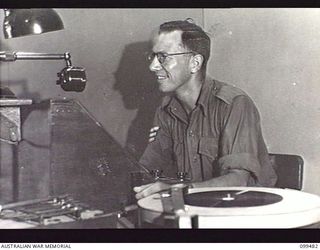 JACQUINOT BAY, NEW BRITAIN, 1945-11-20. ONE OF THE STATION ANNOUNCERS AT HIS TABLE IN THE LARGE STUDIO AT AUSTRALIAN ARMY AMENITIES SERVICE BROADCASTING STATION 9AE