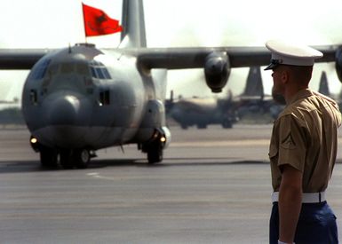 During a Repatriation Ceremony on December 17, 1999, at Hickam Air Force Base, Hawaii, a U.S. Marine salutes as he awaits the arrival of the US Air Force C-130 Hercules cargo plane, in the background, carrying the remains of fallen U.S. Marines from World War II