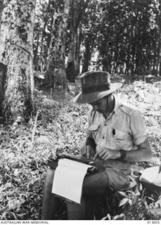 1942-11-19. NEW GUINEA. KOKODA. WAR CORRESPONDENT GEOFF. READING OF THE SYDNEY "DAILY MIRROR" PLUGS AWAY AT A TYPEWRITER IN KOKODA. HIS FEET ARE IN A JAPANESE SLIT TRENCH. (NEGATIVE BY G. SILK)