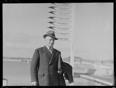 Mr R W Robson, Pacific Islands Publications, standing in front of a sign reading 'Sydney, Singapore, Calcutta, Hong Kong, Alexandria, Durban, London and Empire Flying Boat'