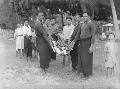 [Full-length group portrait of a two lines of Pacific Island men with root vegetables]