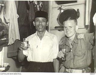 AT SEA. 1945-03-30. CORPORAL D.C. HARDY, CINEMATOGRAPHER, 9 MILITARY HISTORY FIELD TEAM ATTACHED HEADQUARTERS 9 DIVISION, (2), WITH MR. MAT-MOEM, SERANG OF THE VAN HEUTSZ. THE VESSEL CARRIES ..