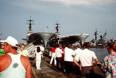 Family members and friends of crew members aboard the amphibious assault ship USS GUAM (LPH-9) watch from the pier as the vessel prepares to depart for the Persian Gulf in response to Iraq's invasion of Kuwait. The amphibious assault ship USS IWO JIMA (LPH-2) is moored to the pier