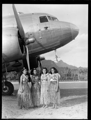 Four unidentified local girls in hula skirts in front of a C47 transport aircraft, Rarotonga airfield, Cook Islands
