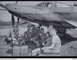 PORT MORESBY, PAPUA, 1942-07-11. UNITED STATES MECHANICS WHO SERVICE THE AIRA-COBRA FIGHTER PLANES WHICH ARE OPERATING IN THE NEW GUINEA WAR ZONE HAVING THEIR MID-DAY MEAL IN THE SHADE OF A WING OF ..