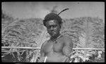 Man with feather hair piece and pipe, near house