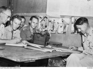 NADZAB, NEW GUINEA. C. 1944-02. MEMBERS OF NO. 24 SQUADRON RAAF WORK AT A TABLE IN THE OPERATIONS ROOM. NOTE THE POSTERS OF TIVOLI GIRL PERFORMERS FROM THE TIVOLI THEATRE ON THE WALL IN THE ..