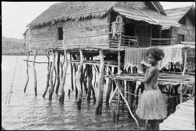 Woman wearing a grass skirt with nets drying and village houses behind, Hanuabada, Papua, 1921, 2 / Sarah Chinnery