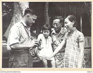 BITAPAKA, NEW GUINEA. 1945-11-04. MR H. MEEN CHIN, AGRICULTURAL DEPARTMENT CLERK PHOTOGRAPHER, SHOWING HIS CAMERA TO MR T. MOW, HIS WIFE AND BABY GIRL. MR MOW WAS A SCHOOL TEACHER AT THE CHINESE ..