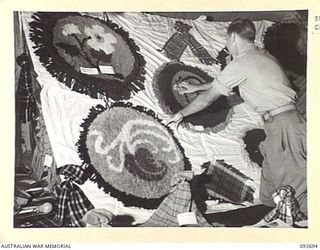 LAE AREA, NEW GUINEA. 1945-06-03. DURING AN EXHIBITION OF HOBBIES AND CRAFTS CORPORAL G.S. BOON, AN INSTRUCTOR OF ARTS AND CRAFTS AT 112 CONVALESCENT DEPOT, IS PICTURED AMONG AN ARRAY OF WOOLLEN ..