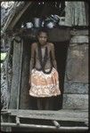 Momwarka's mother, wearing black mourning necklace, stands in doorway of small house, kitchen items above her