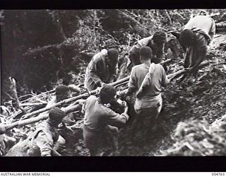 BULLDOG ROAD, NEW GUINEA. 1943-07-19. NATIVES CARRYING COMPRESSOR PARTS UP A SLIPPERY, MUDDY TRACK AT ECCLESTONE GAP