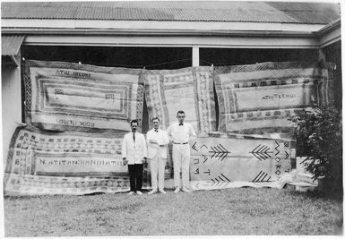 Unidentified men in front of a display of atiu mats, Cook Islands