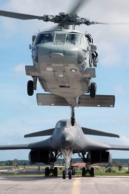 A US Navy (USN) MH-60S Seahawk helicopter, Helicopter Sea Combat Squadron 25 (HSC-25), Island Knights, maneuvers over the taxiway at Andersen Air Force Base (AFB) on the US territory of Guam. Behind the Seahawk is a US Air Force (USAF) B-1B Lancer bomber, 34th Bomb Squadron (BS), Ellsworth AFB, South Dakota (SD). HSC-25 is a permanently forward-deployed squadron, homeported on board Andersen AFB