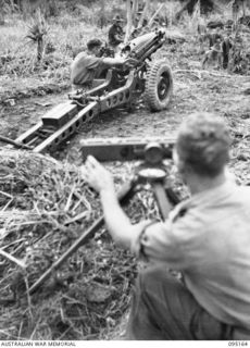 KIARIVU, NEW GUINEA, 1945-08-11. SERGEANT G. MCGOVERN (2), INDICATING THE ADJUSTMENT TO THE GUNNER ON THE PARALLELOSCOPE AS GUNNER A.P. GREEN (1), PREPARES THE 75MM GUN FOR FIRING. THE MEN ARE ..