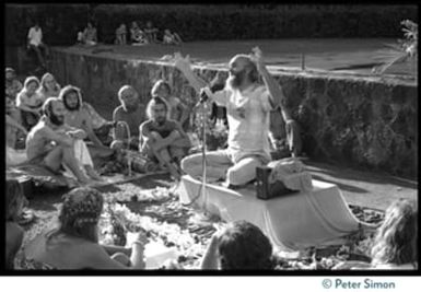 Ram Dass seated on a small platform, arms raised, during his appearance at Andrews Amphitheater, University of Hawaii