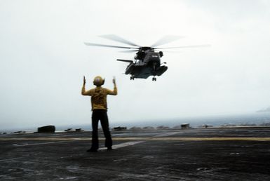 A plane director signals landing instructions to the pilot of a Helicopter Mine Countermeasures Squadron 14 (HM-14) RH-53D Sea Stallion helicopter hovering over the flight deck of the amphibious assault ship USS GUAM (LPH-9)