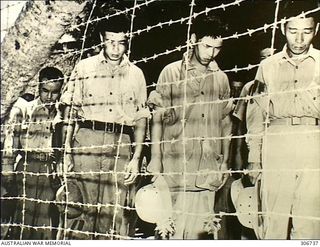 Guam. 1945-08-15. Japanese prisoners of war (POW), bow their heads and remove their hats in reverence for Emperor Hirohito, as they stand behind a barbed wire fence of a POW camp, to hear the radio ..
